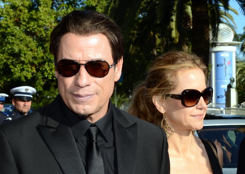 John Travolta and Kelly Preston walking together at Cannes, both wearing dark clothing and large sunglasses on a sunny day.