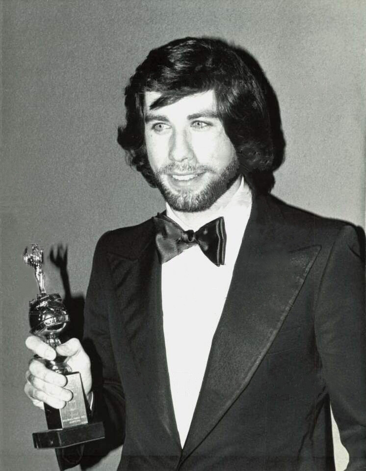 A young bearded John Travolta in a black tuxedo and bow tie holding a Golden Globe statuette, photographed in black and white.