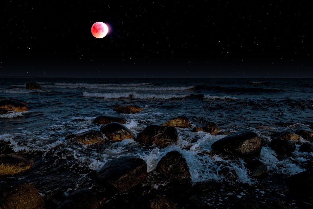 A blood moon tinged red and pink hangs in a starry night sky above a rocky coastline, with ocean waves crashing over dark boulders in the foreground.