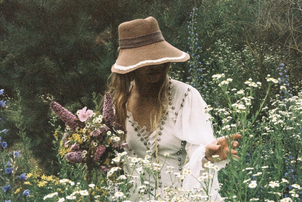 Woman in a white embroidered dress and woven sun hat picks wildflowers in a sunlit garden.