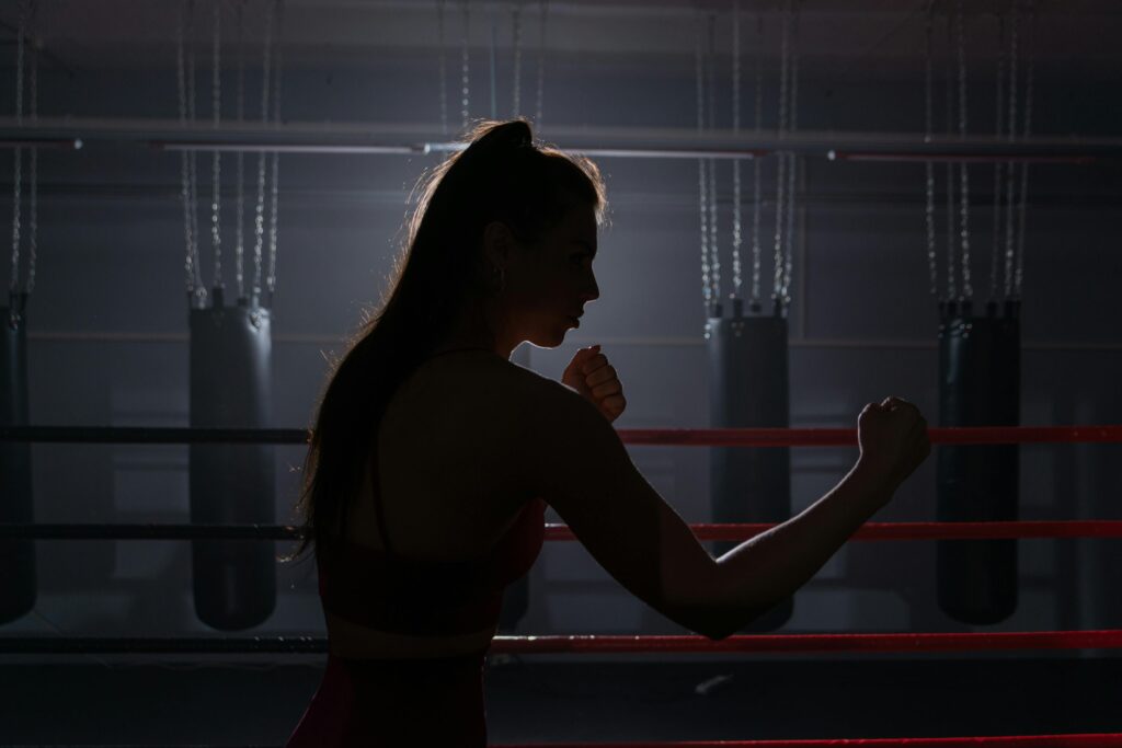 Silhouette of a woman in a boxing stance, backlit in a boxing ring with lights hanging overhead.