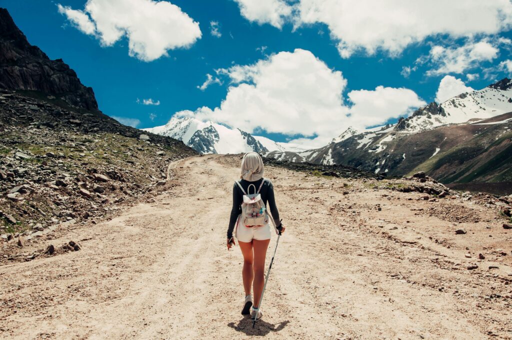 Woman with a backpack and hiking poles walks alone down a dusty mountain path toward snow-capped peaks under a blue sky.