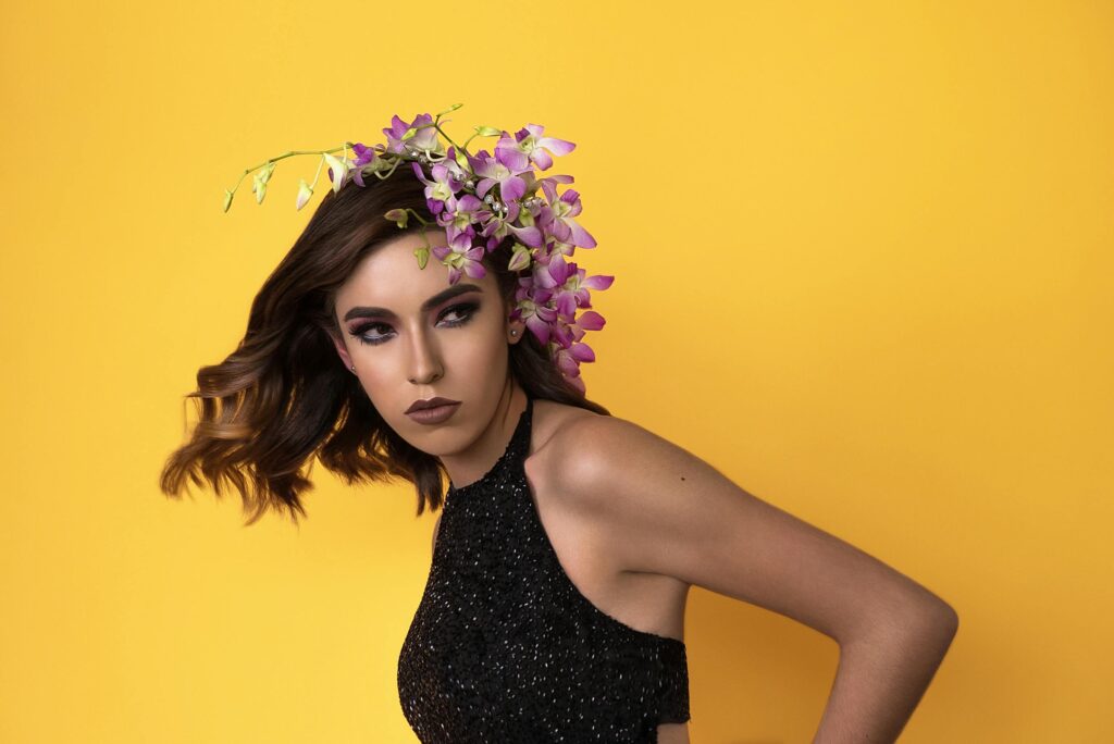 Woman in a black sparkly halter top poses against a bright yellow background, purple orchids woven through her wavy brown hair.
