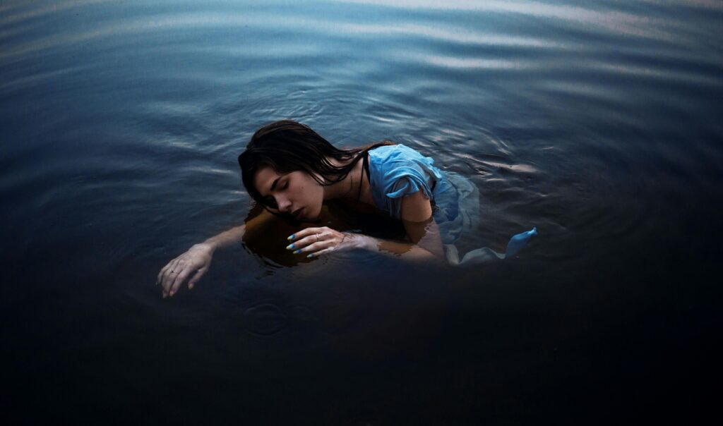 Woman in a light blue dress rests her head on her arm in dark water, eyes closed, her body curled as ripples move gently around her.