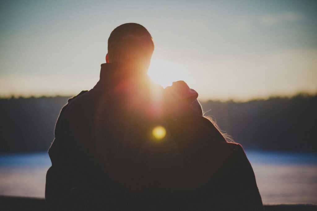 Silhouette of a couple embracing from behind as golden sunlight flares between them, a lake and trees in the background.