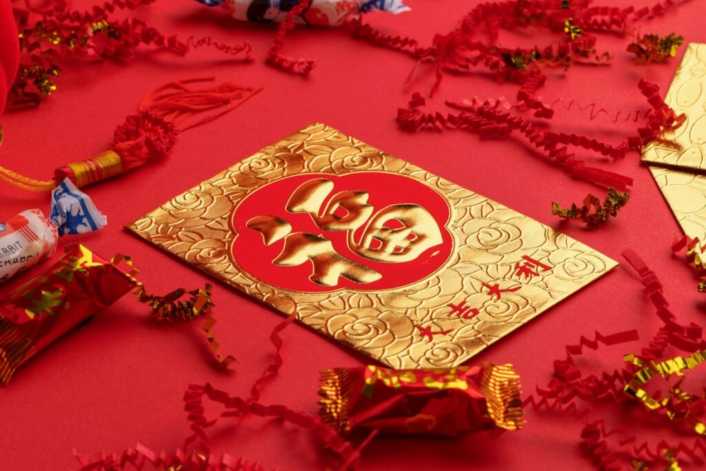 A gold and red Chinese New Year envelope with an embossed lucky character design, surrounded by wrapped candies and red and gold streamers on a bright red background.