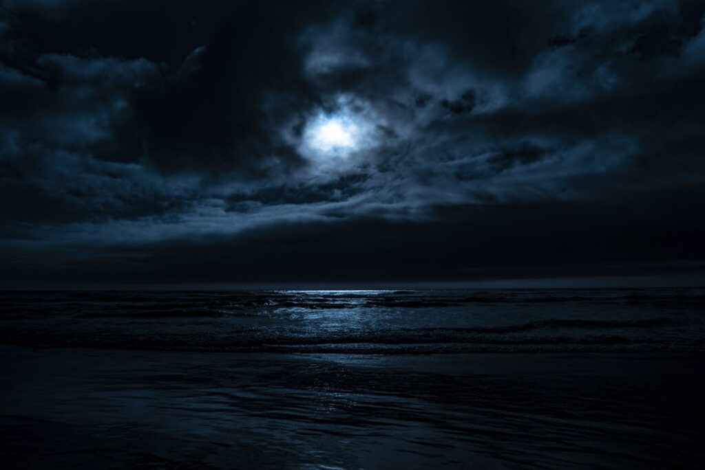 A dark ocean at night with heavy clouds covering most of the sky, broken only by a single patch of moonlight pushing through near the center. The moon casts a pale blue-white glow across the water below it, catching the surface of the waves in a narrow strip of light while the rest of the sea and shoreline disappear into near-total darkness.