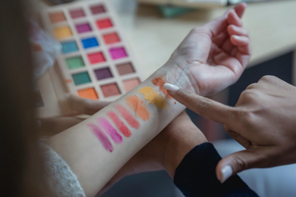 Makeup artist swatching eyeshadow colors on a person's inner forearm, with orange, pink, and magenta stripes visible alongside a colorful eyeshadow palette.
