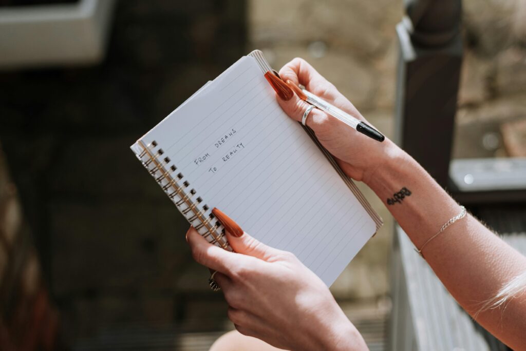 A person holding a small spiral-bound notebook open with "from dreams to reality" handwritten in lowercase on the lined page. They hold a black pen in the other hand, resting against the page. Their nails are long and painted orange-red, and they're wearing silver rings on both hands. A word tattoo is visible on the inside of their wrist, and a thin silver bracelet sits just below it. The background is blurred and outdoor.