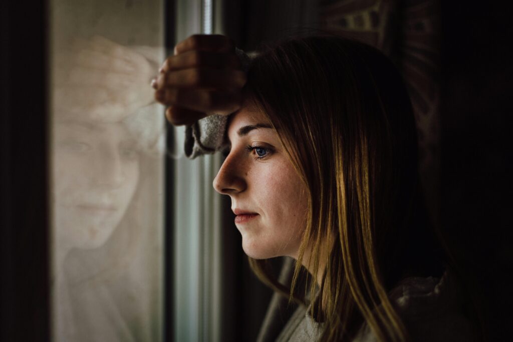 Woman gazes pensively out a rain-speckled window, her reflection faintly visible in the glass.