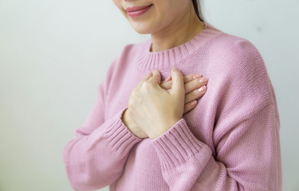 A woman in a soft pink knit sweater pressing both hands gently against her upper chest, fingers overlapping. Her face is cropped above the mouth showing a slight smile. The background is plain and light-colored, and the framing is tight on her hands and torso.
