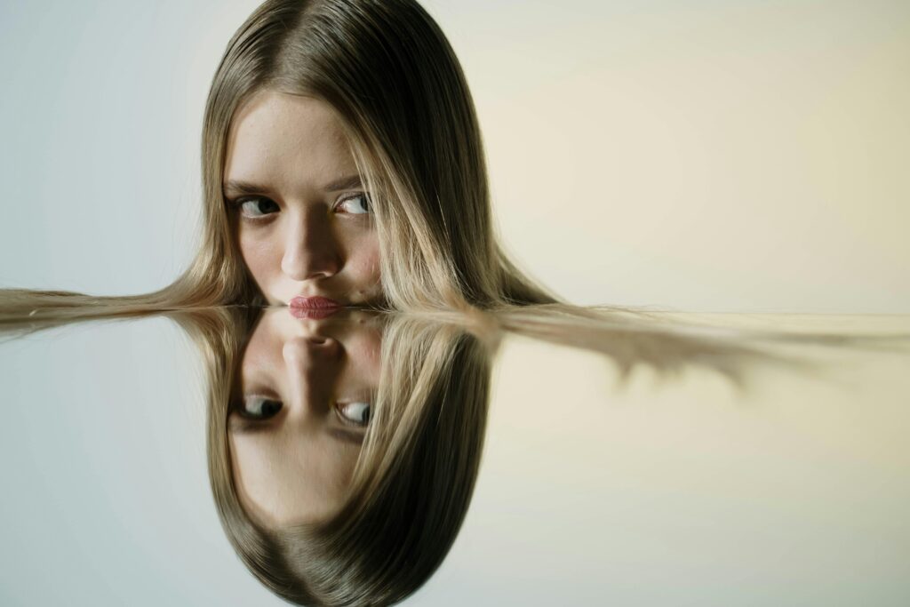 A young woman with long straight blonde hair resting her chin on a reflective surface that mirrors her face and hair symmetrically. She looks slightly off-camera with a calm, searching expression. The background is warm and neutral, and her hair extends horizontally across both the real and reflected image creating a continuous line.