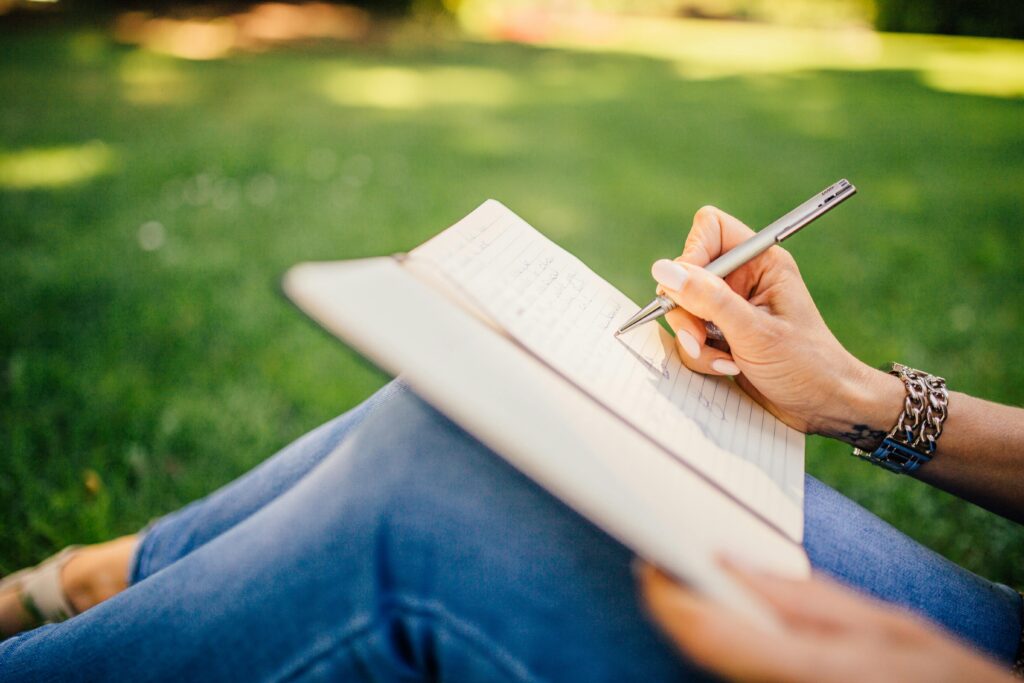 A person sitting cross-legged on green grass wearing blue jeans, writing with a silver pen in an open lined notebook balanced on their lap, their wrist adorned with layered bracelets.