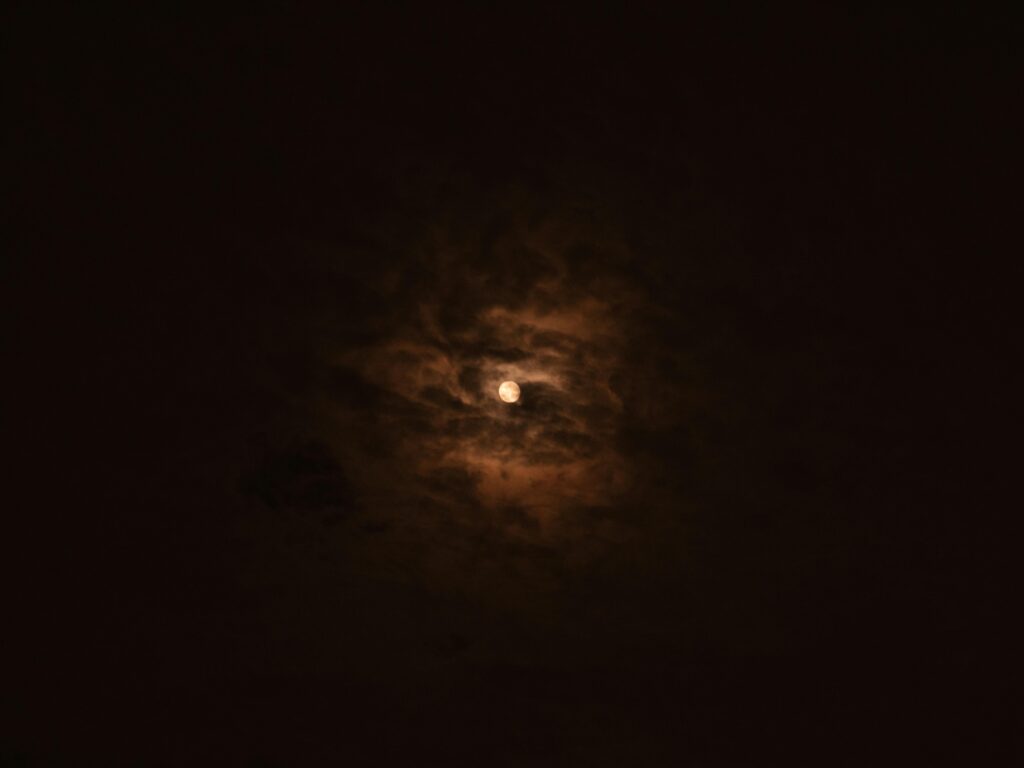 A small bright moon partially obscured by thin, wispy clouds that catch its light in warm brown and amber tones, surrounded by near-total darkness.