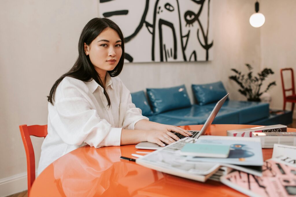 Woman in a white button-down shirt works at a laptop in a modern space with a blue sofa and abstract art behind her.
