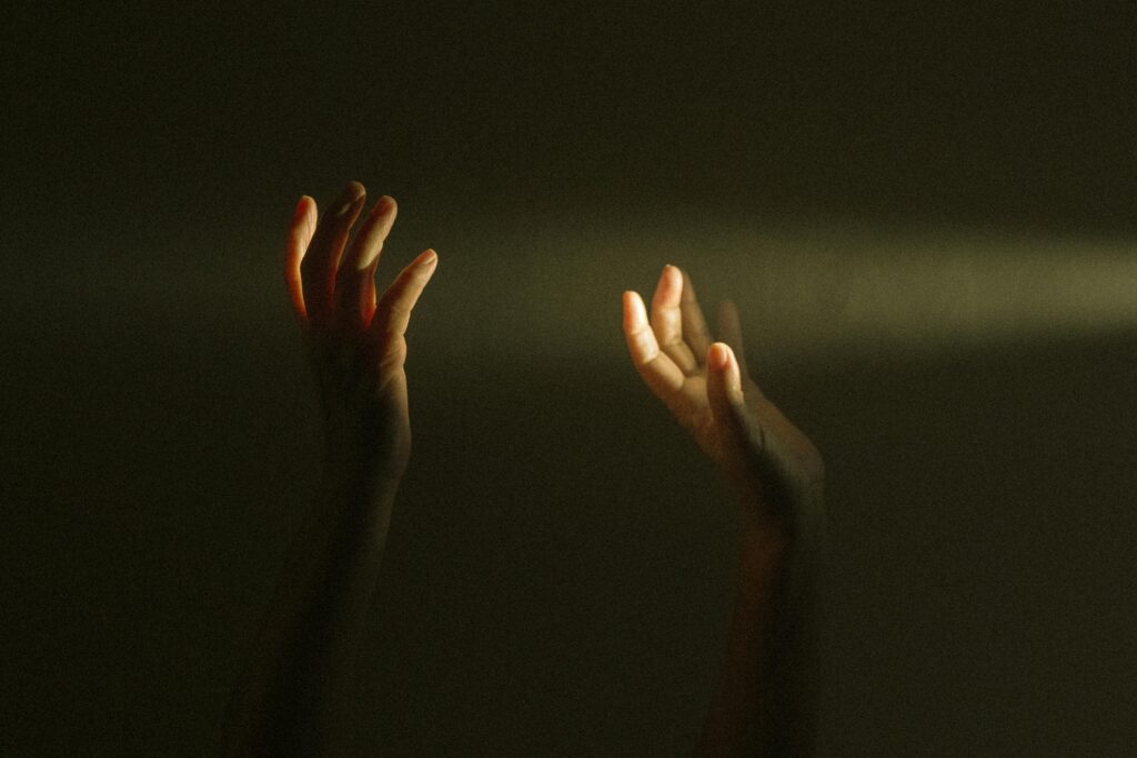 Two hands reach upward into a beam of light against a dark, shadowy background.