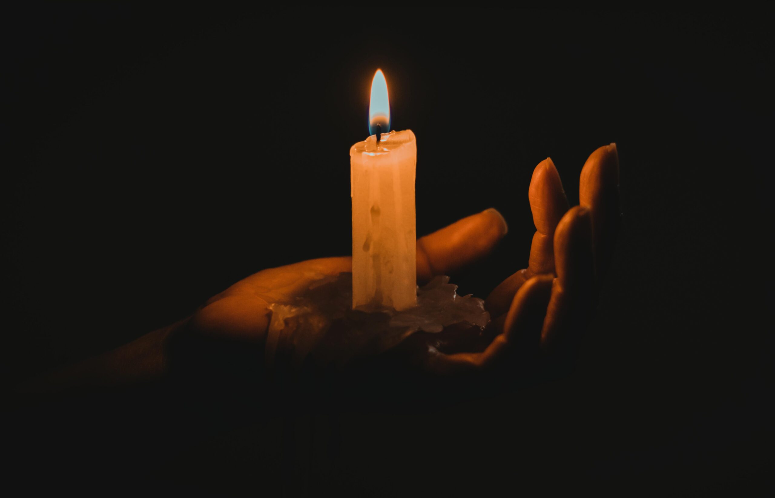A hand emerges from darkness holding a lit candle, the warm flame illuminating only the fingers and wax.