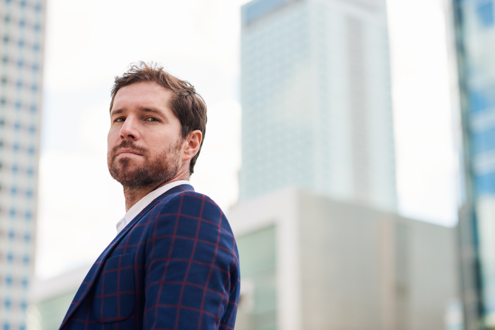 Confident and successful young businessman wearing a blazer standing alone in the city with office buildings in the background