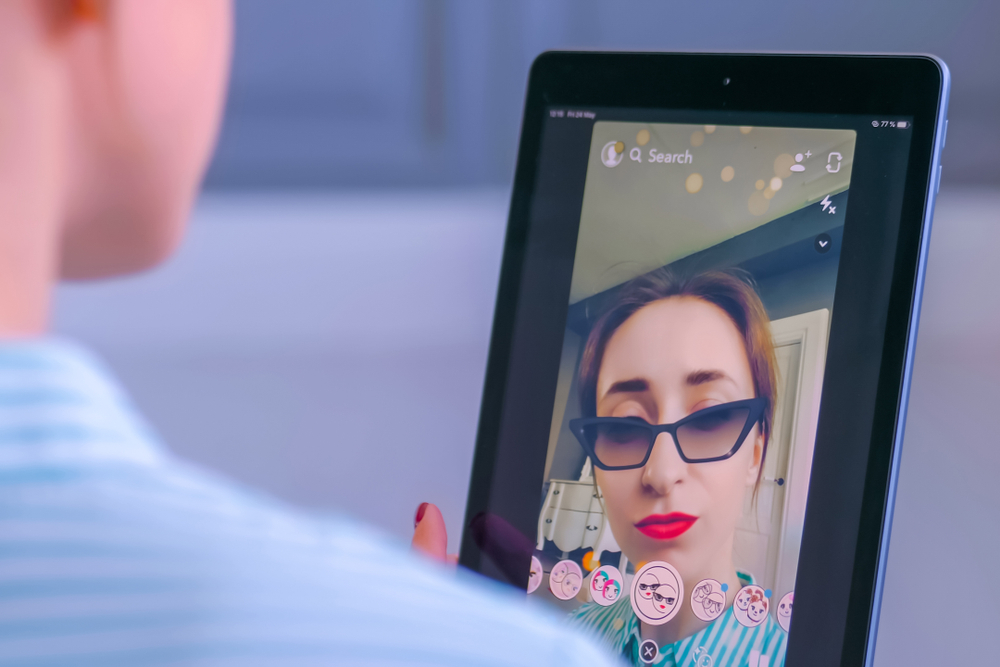 MOSCOW, RUSSIA - MAY 24, 2019: Woman using Snapchat multimedia messaging app with 3d face mask filter on tablet at home. Face detection, AR, beauty, selfie, vogue, fashion, trendy concept