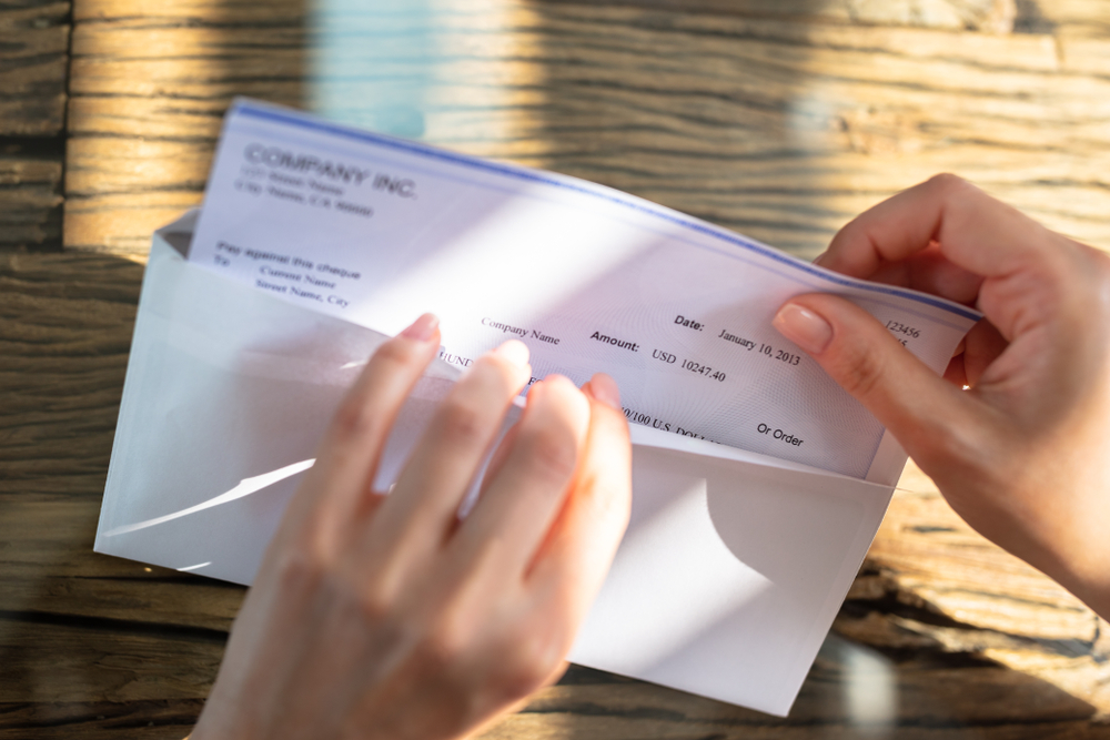 Close-up Of A Businessperson's Hand Opening Envelope With Paycheck Over Wooden Desk