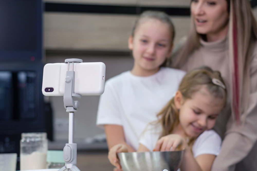 Mom teaches her daughters to cook dough in the kitchen. The family is filming a culinary video on a smartphone