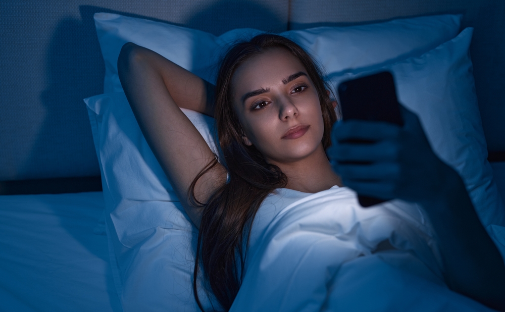 From above young female with hand behind head lying on soft pillow on bed and browsing social media, on smartphone while suffering from insomnia at night at home