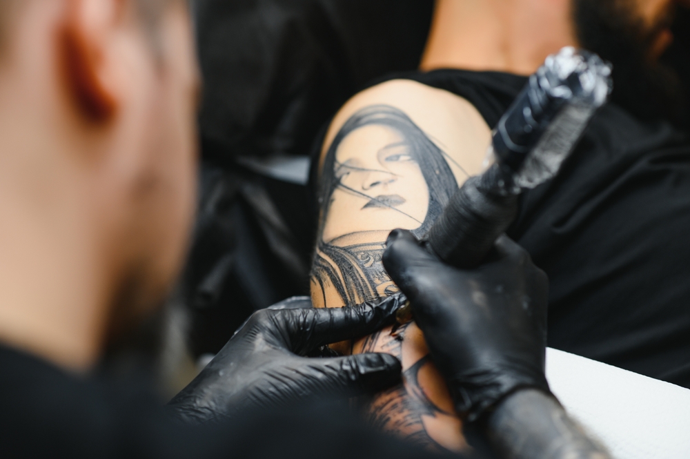 Cropped close up of a bearded tattoo artist working at his studio tattooing sleeve on the arm of his male client. Man getting tattooed by professional tattooist