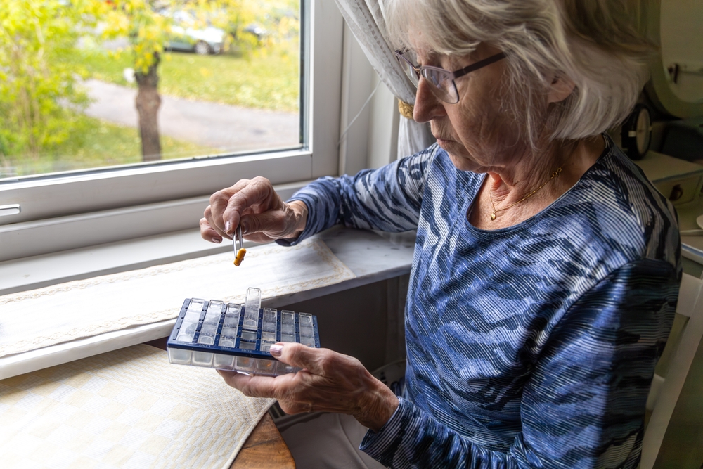 An elderly, gray-haired woman meticulously organizes her medicine pills using tweezers, seated by the window in the comfort of her own home.