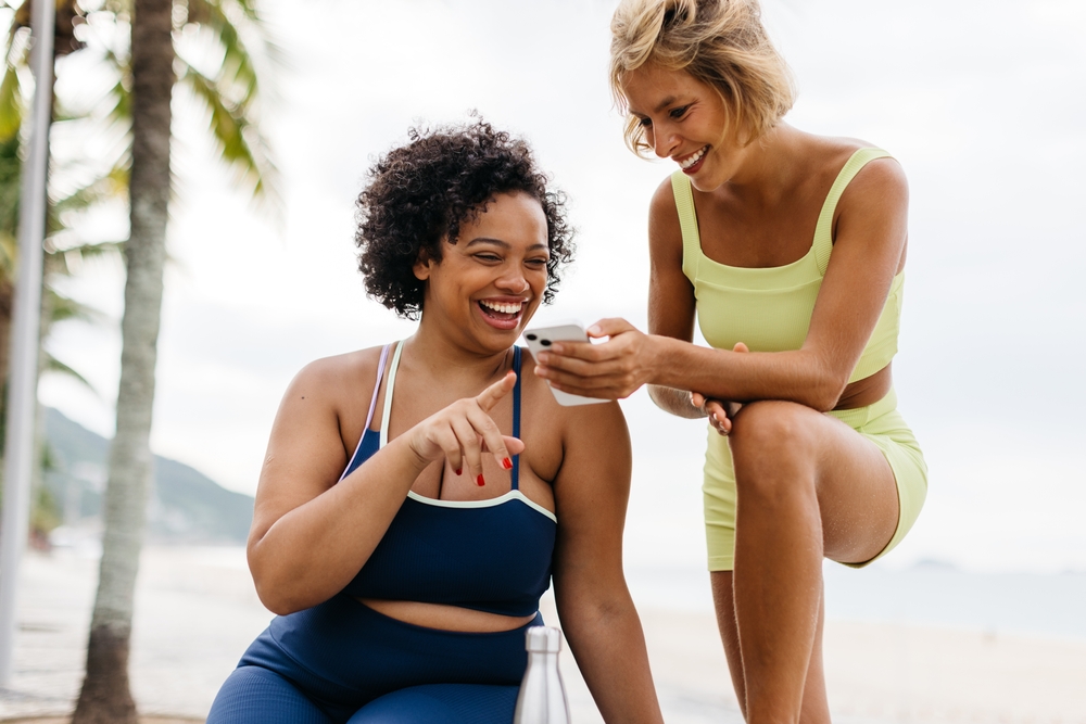 Two fit women taking a moment to browse social media on their workout break near the beach. Happy fitness buddies enjoying their outdoor exercise session.