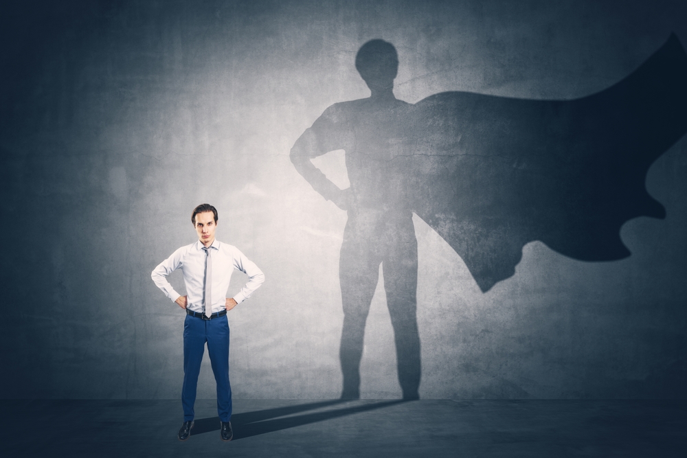 Attractive young european businessman standing on concrete wall background with cape shadow. Leadership, success and power concept
