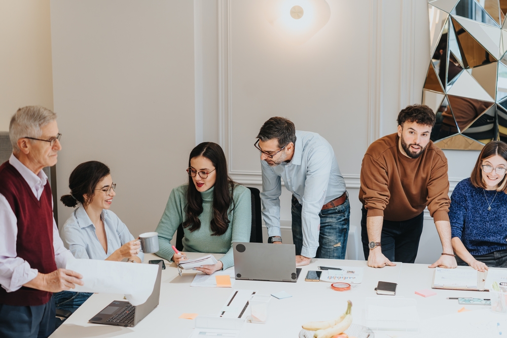A multigenerational group of men and women gather around a table, collaborating in a business meeting focused on strategy and innovation.