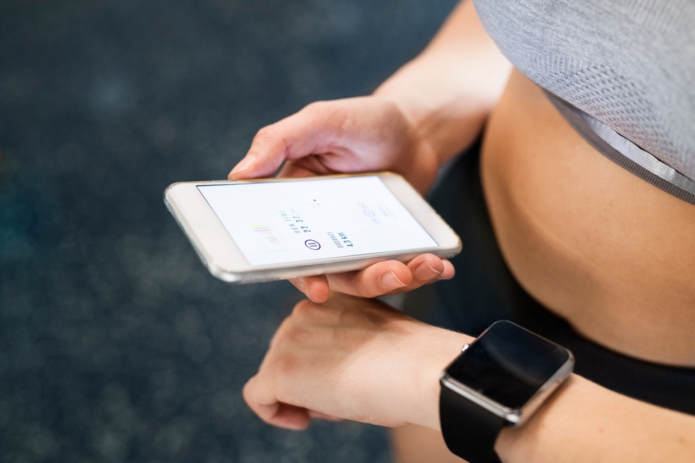 Young fitness girl checking her performance on smatphone and smartwatch after hard workout in gym.