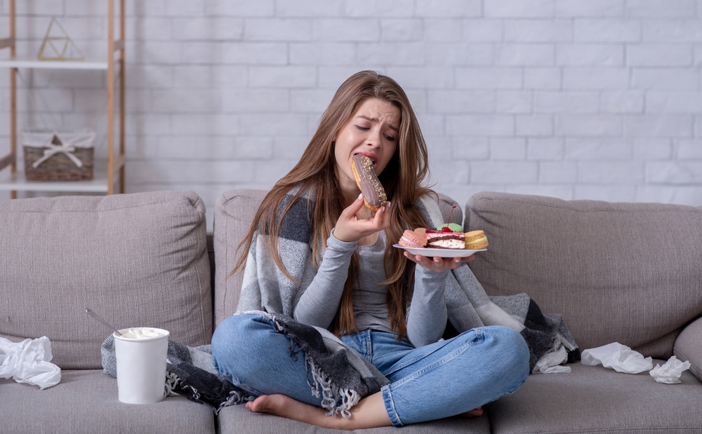 Comfort foods and stress eating concept. Frustrated young lady devouring pastry on sofa at home. Upset woman dealing with negative emotions, anxiety or problems through sweets