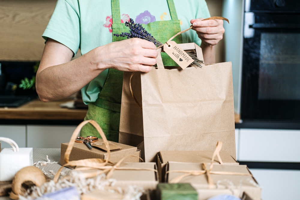 A small business owner ties a Handmade with love tag and lavender sprig to a brown paper bag surrounded by gift boxes. Home-based business, handmade goods. High quality photo