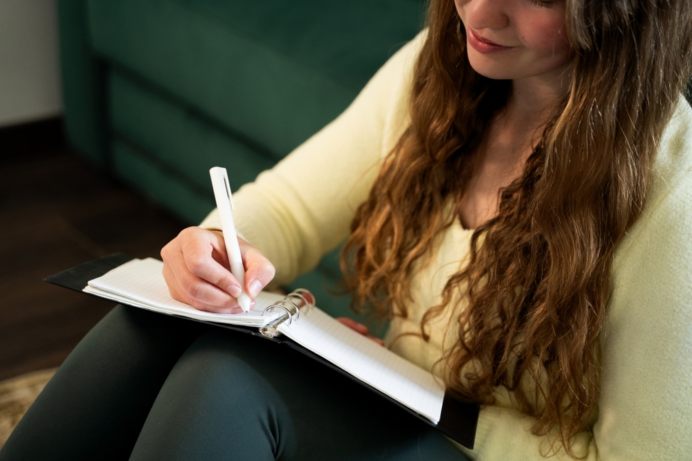 Smiling young woman sitting on floor at home and writing in notebook journaling taking notes in diary, creative process, self reflection, remote education, casual lifestyle, mental health