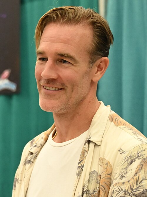 James Van Der Beek smiling, photographed from the chest up against a teal curtain backdrop. He has short sandy-blond hair and light stubble, and he's wearing an open floral-print button-up shirt over a white crewneck tee.