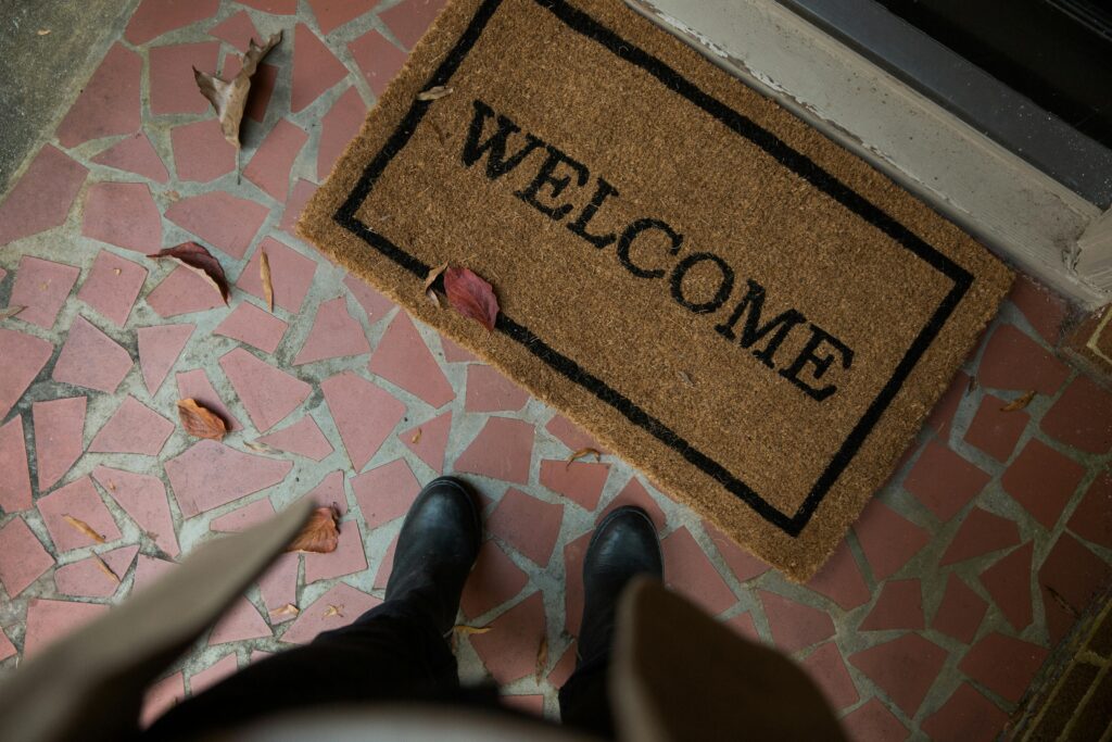 A person in black boots standing on a pink mosaic porch in front of a woven welcome mat, with dried autumn leaves scattered across the tiles.