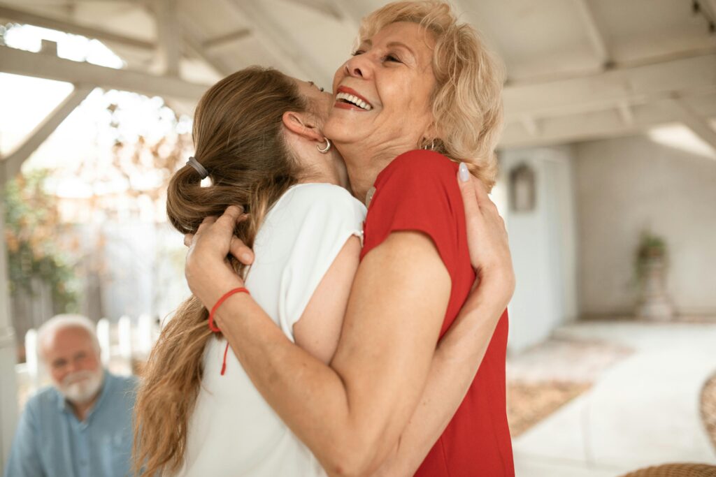 An older woman in a red top laughing as she hugs a younger woman with a long ponytail, while an older man with a white beard watches from the background of a sunlit porch.