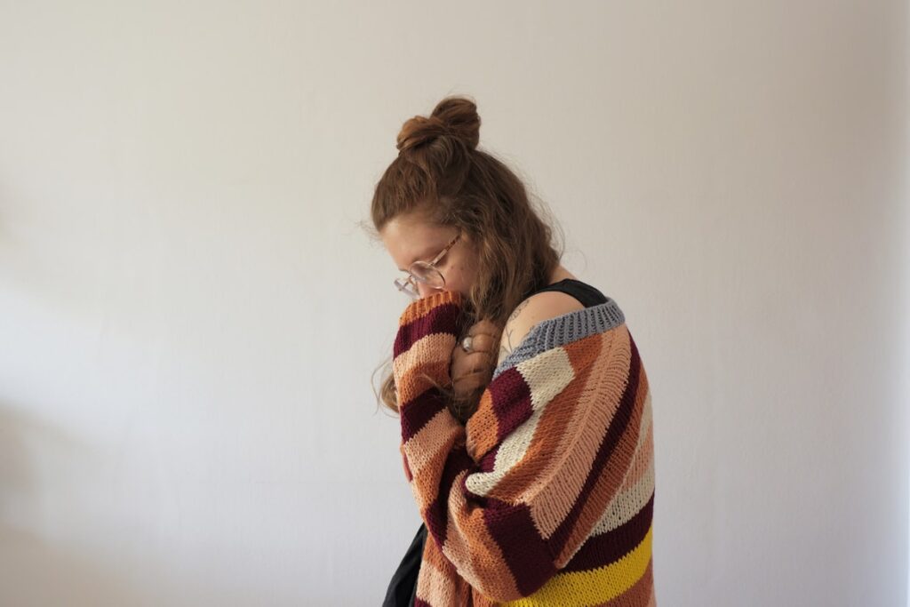 Woman with curly hair in a messy bun wears glasses and a chunky striped knit cardigan in burgundy, orange, cream, and grey. She holds the collar up to her nose, smelling the fabric, against a plain wall.