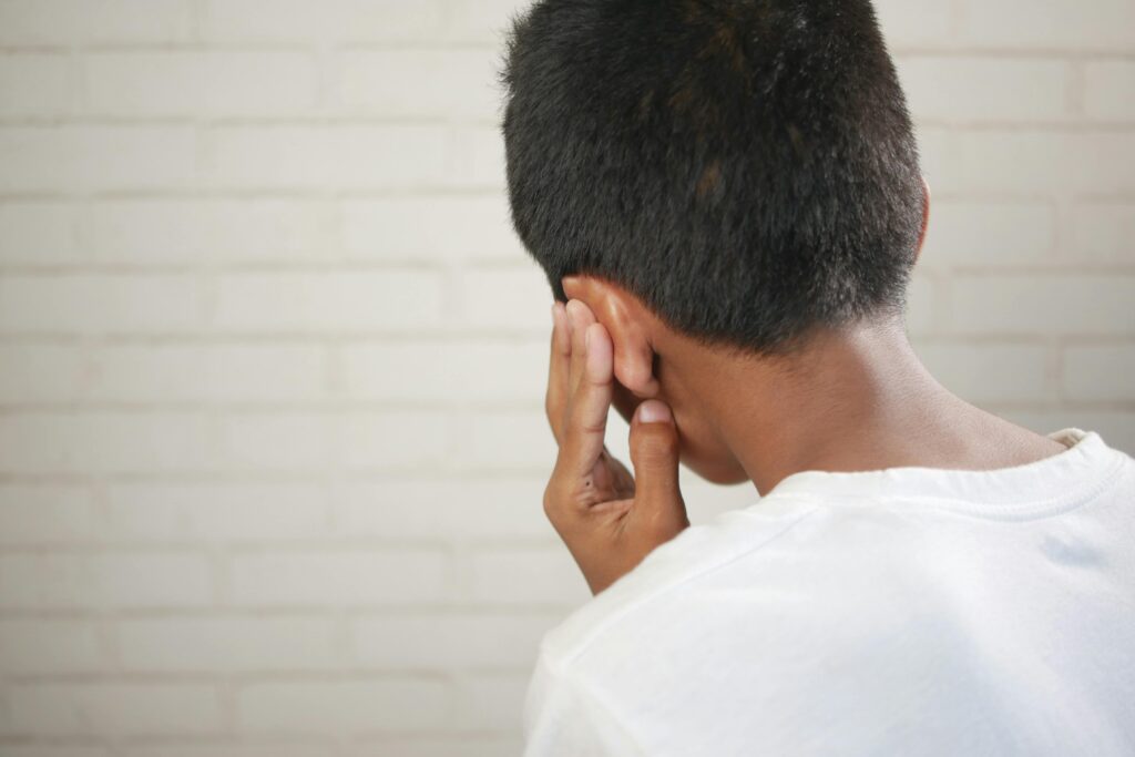 Back view of a person with short dark hair wearing a white t-shirt, pressing their hand against the side of their head near their ear, suggesting discomfort or a headache.