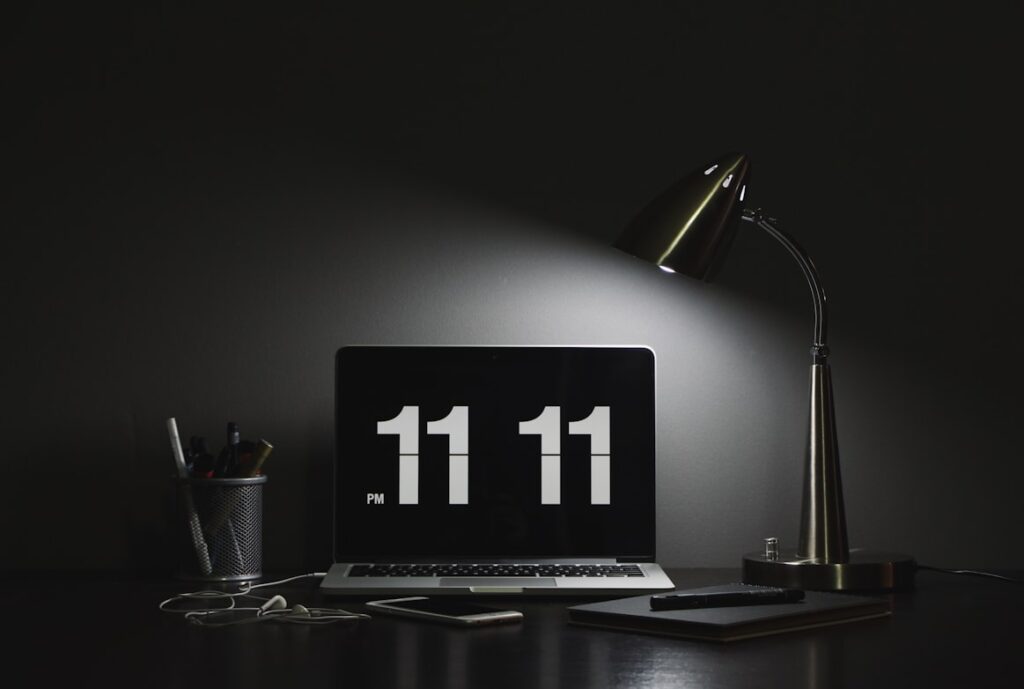 Minimalist dark desk setup with a laptop displaying "11:11 PM" in large flip-clock numerals. A metallic desk lamp provides the only light, illuminating a pencil holder nearby.