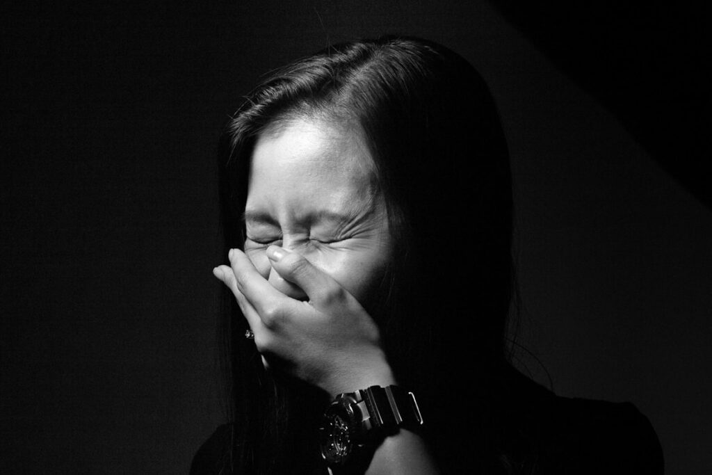 Black and white portrait of an Asian woman with long dark hair sneezing, her hand covering her mouth and nose, eyes squeezed shut.