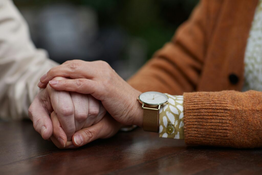 Two older hands clasped together on a dark wooden table, one wearing a gold watch with a patterned sleeve, the other in a light-colored top, with soft green foliage blurred in the background.