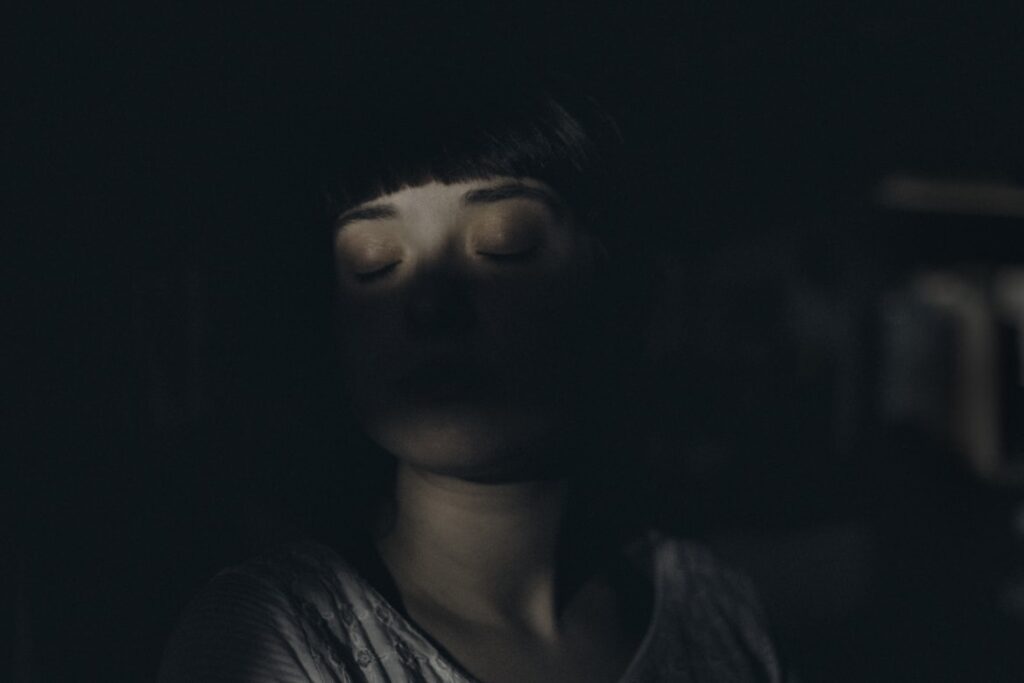 Dark artistic portrait of a person with short dark hair and bangs, eyes closed. Heavy shadow obscures the lower half of their face, creating a moody, atmospheric effect.