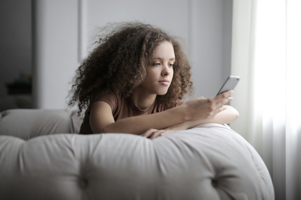 Young woman with voluminous curly dark hair lies on a light grey sofa, propped on a pillow, looking at her smartphone with a contemplative expression.