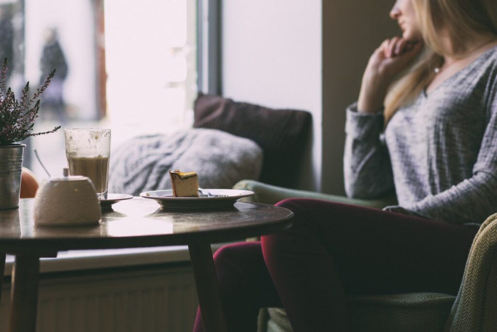 Woman with blonde hair in a grey sweater sits in a burgundy velvet cafe chair, gazing toward a window. A small round table holds a layered latte in a tall glass, a slice of cheesecake, and a vase of dried heather. Soft natural light fills the cozy space.