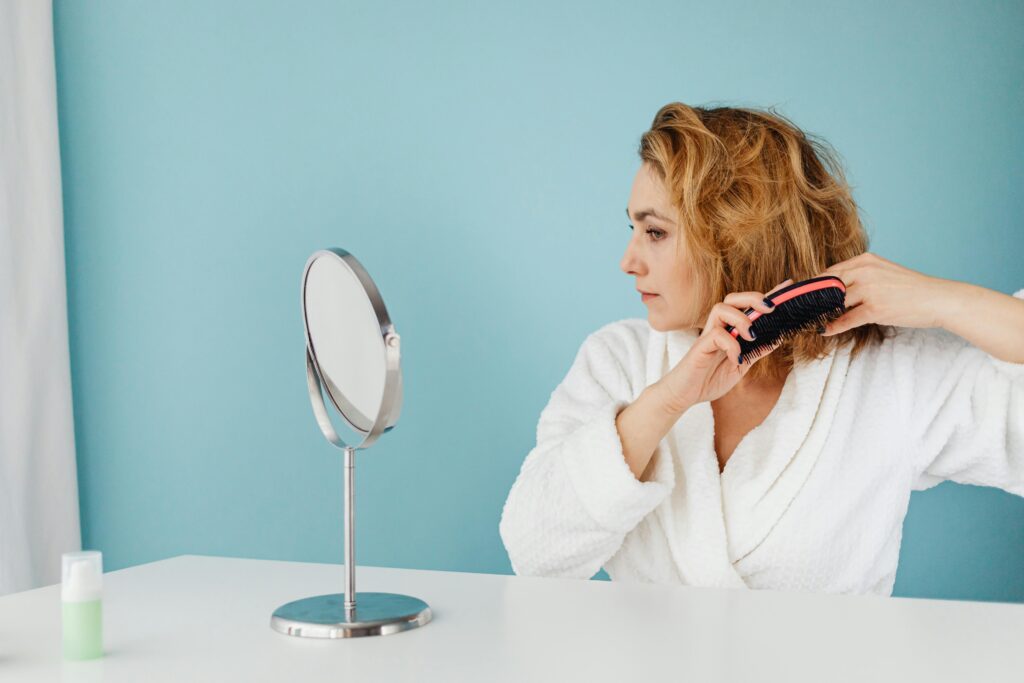 Woman in a white bathrobe brushing her short curly hair while looking into a round vanity mirror against a light blue background.