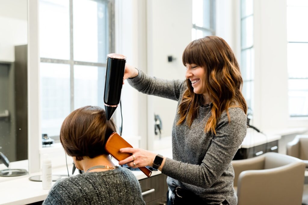 Hairstylist blow-drying a client's short bob haircut in a bright salon, using a round brush.