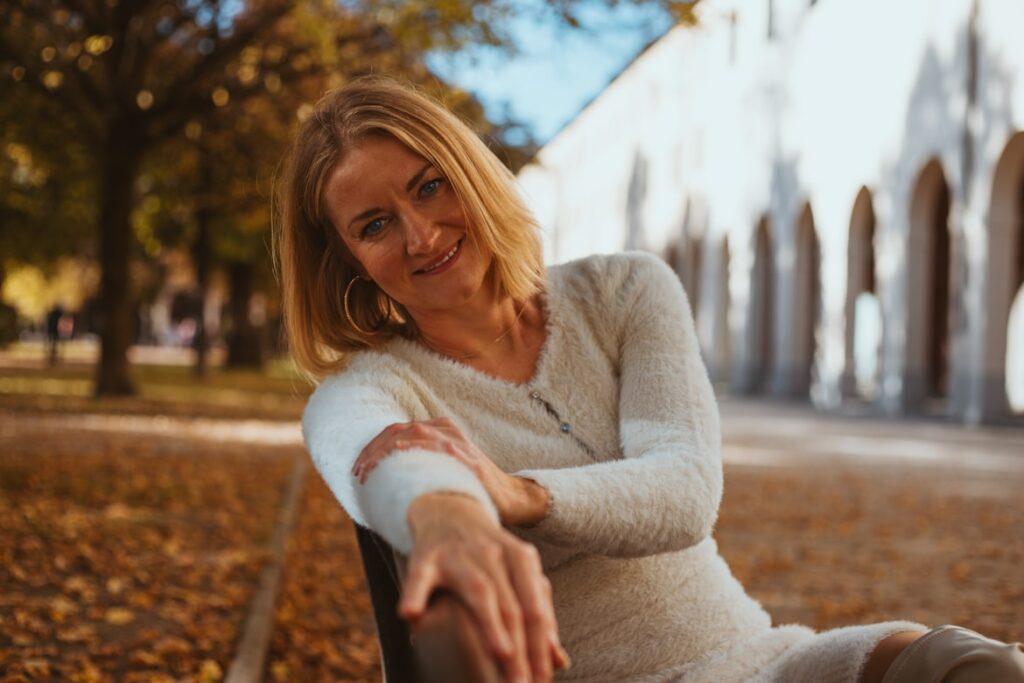 Woman with shoulder-length blonde hair in a white sweater, sitting on a bench surrounded by autumn leaves.