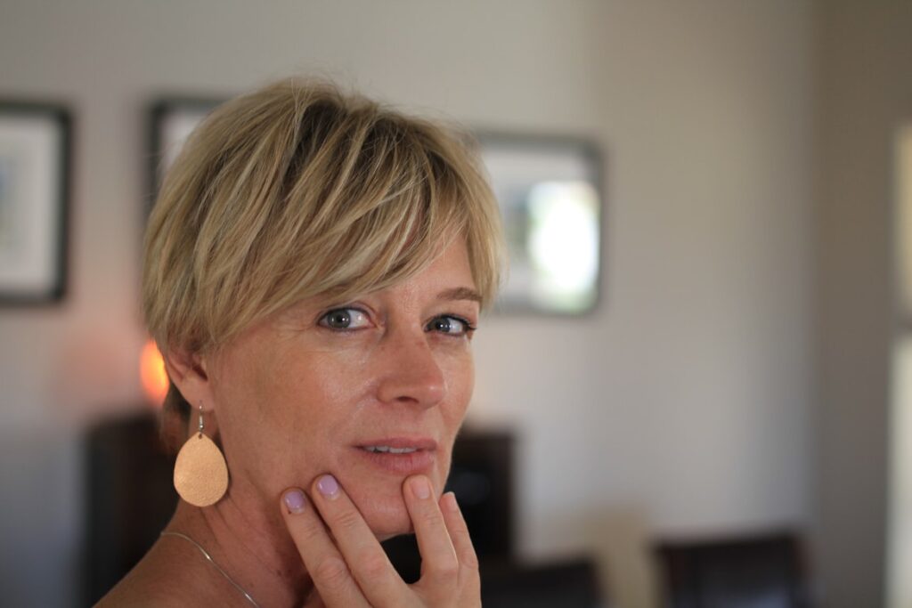 Woman with a short blonde pixie cut and teardrop earrings, resting her chin on her hand in a thoughtful pose.